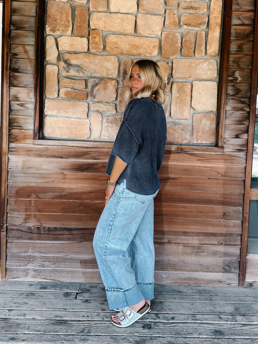 A woman stands on a wooden deck in front of a rustic stone and wood wall, wearing a navy ribbed knit top, light blue wide-leg jeans, and white slide sandals.