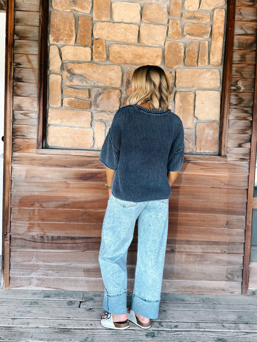 A woman stands with her back to the camera wearing a dark charcoal gray ribbed knit top and light blue wide-leg jeans, posing in front of a rustic stone and wood wall.