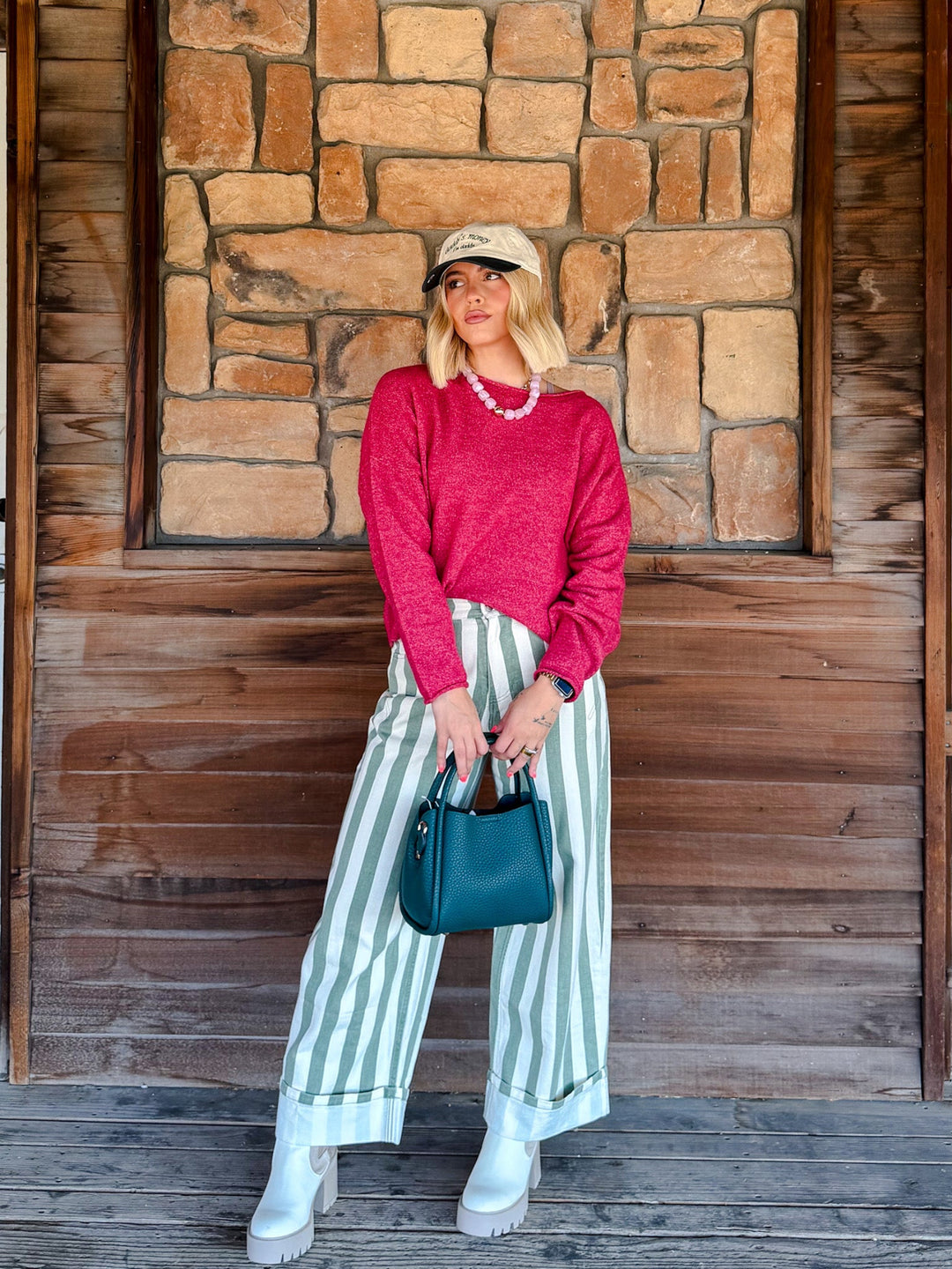 A woman stands in front of a rustic stone and wood wall, wearing a bright pink knit sweater, green and white vertically striped wide-leg pants, white platform boots, a beige baseball cap, and holding a teal handbag.