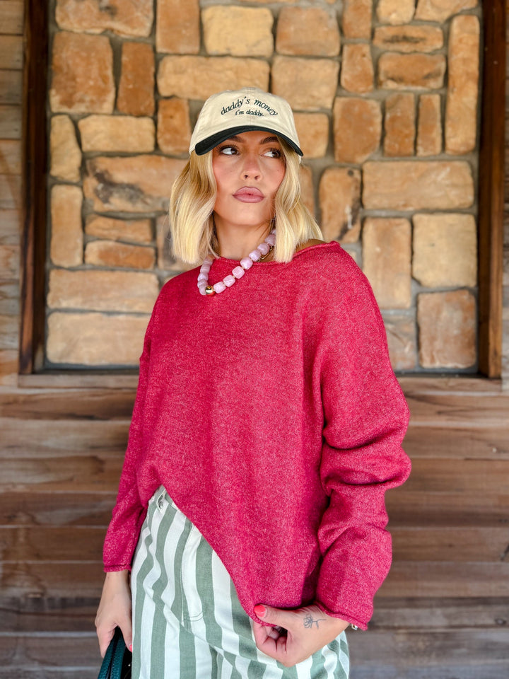 A woman wearing a bright pinkish-red heathered knit sweater, a beige baseball cap, a pink beaded necklace, and green and white striped pants, standing in front of a rustic stone wall.