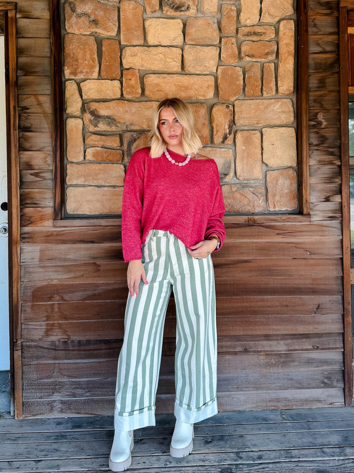 A woman stands on a wooden porch in front of a stone wall, wearing a bright pink cropped long-sleeve sweater, sage green and white vertically striped wide-leg jeans, and white boots.