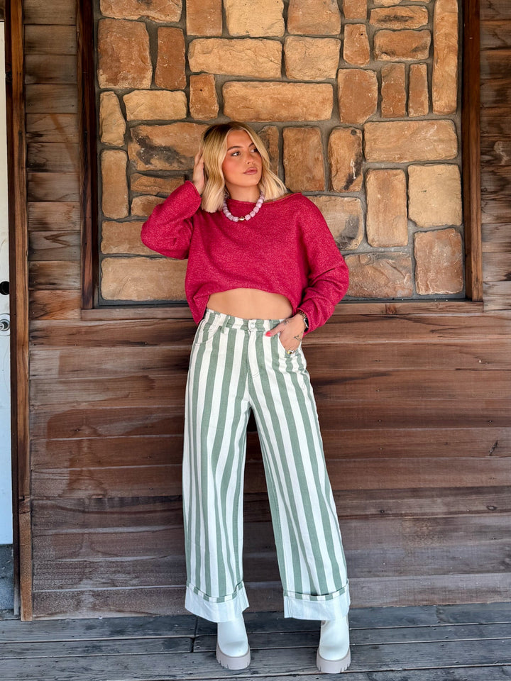 A woman stands on a wooden porch in front of a stone wall, wearing a bright pink cropped long-sleeve sweater, sage green and white vertically striped wide-leg jeans, and white boots.