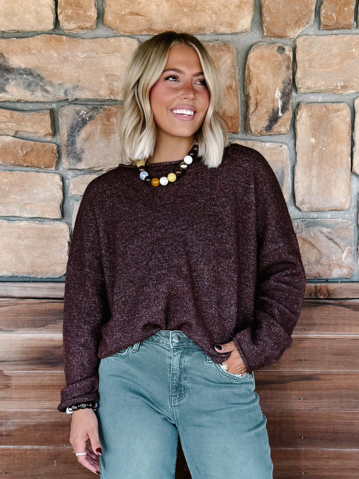 A woman with blonde hair wearing a dark plum-brown heathered knit sweater and light blue jeans, standing in front of a rustic stone wall.
