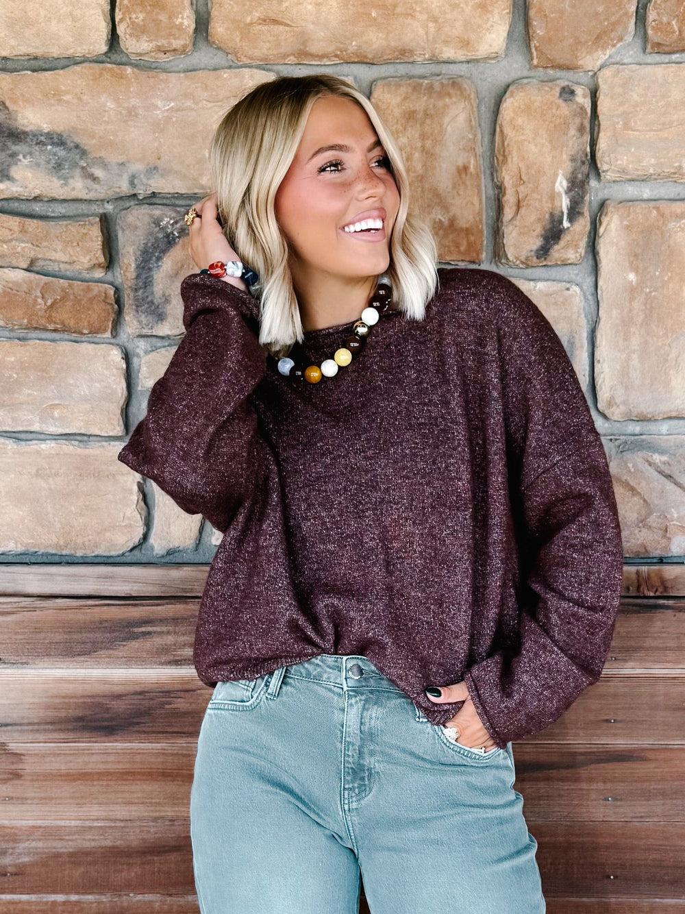 A woman with blonde hair wearing a fuzzy dark plum-brown long-sleeve knit sweater and light blue jeans, posing in front of a rustic stone wall and wooden bench.