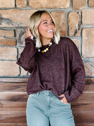 A woman with blonde hair wearing a fuzzy dark plum-brown long-sleeve knit sweater and light blue jeans, posing in front of a rustic stone wall and wooden bench.