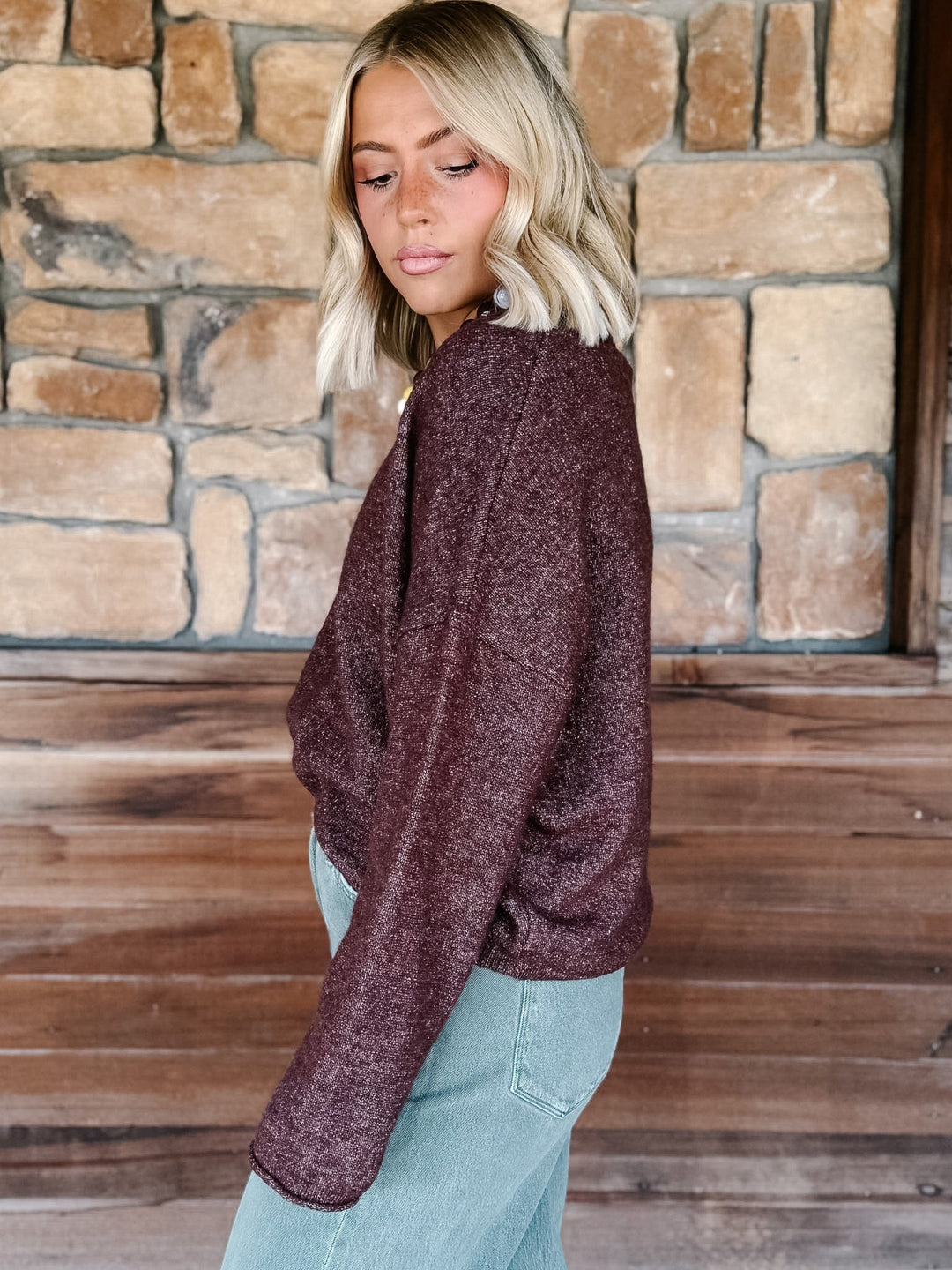 A woman with blonde wavy hair wearing a fuzzy, textured dark plum-brown long-sleeve crop sweater and light blue jeans, standing in front of a rustic stone wall and wooden door.