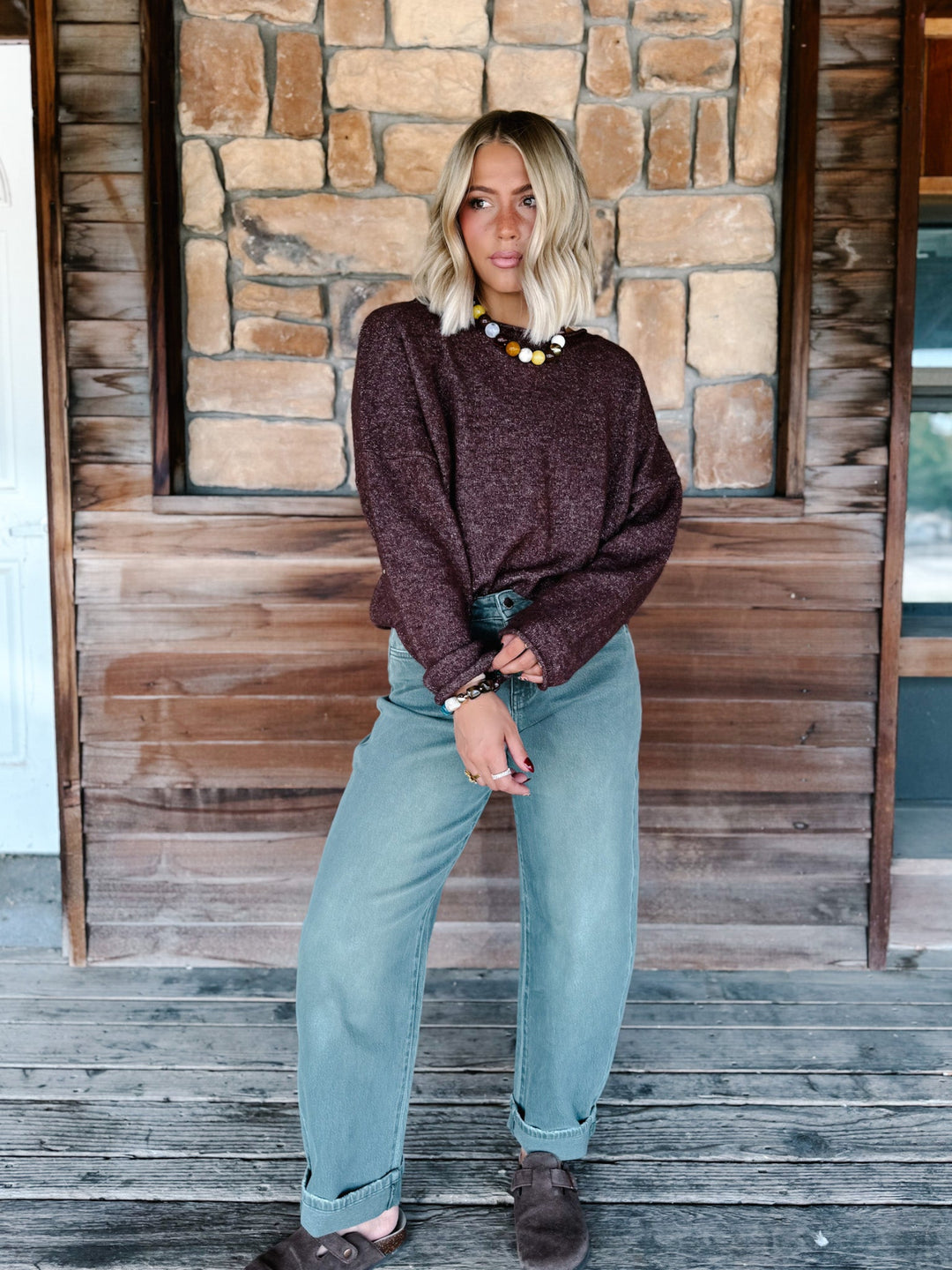 A woman stands on a wooden porch wearing a dark brown heathered knit sweater, light blue wide-leg jeans, and brown shoes, with a chunky multi-colored beaded necklace.