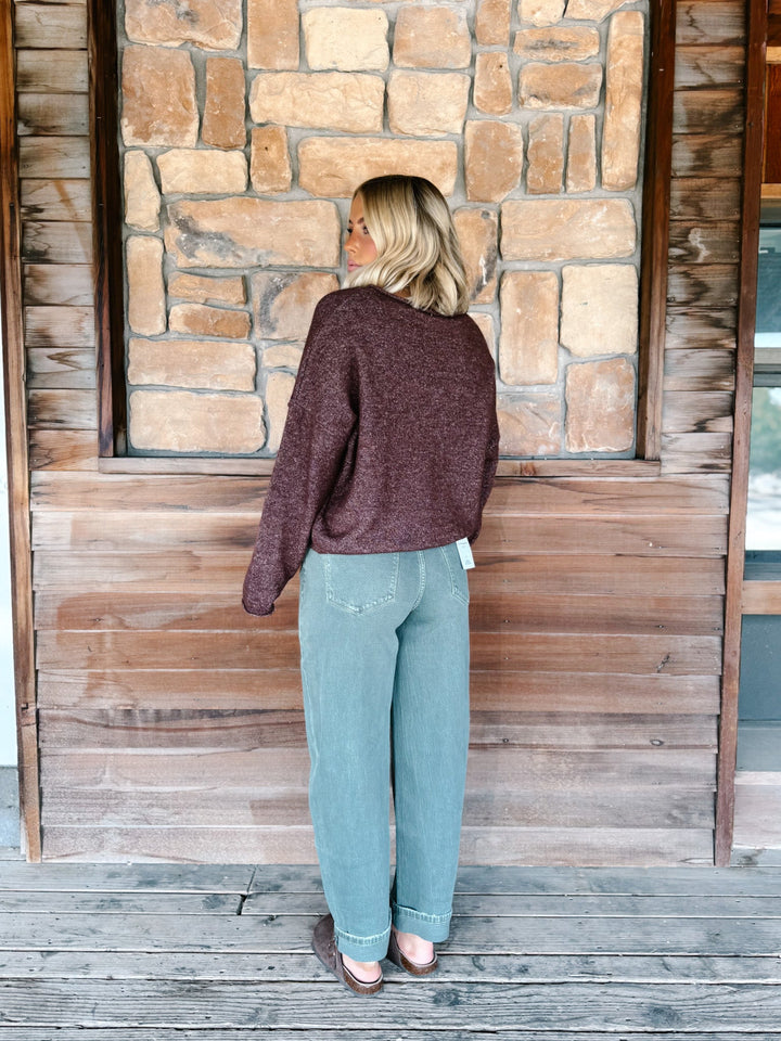 A woman stands on a wooden deck in front of a stone and wood wall, viewed from behind. She is wearing a dark heathered burgundy knit sweater and light blue straight-leg jeans.