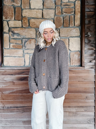 A woman wearing a chunky gray cable-knit cardigan with brown buttons, a white knit beanie, and white pants, standing in front of a rustic stone and wood wall.