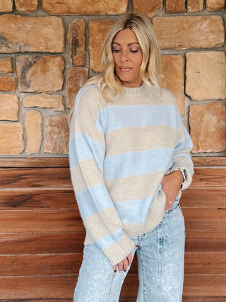 A woman stands on a wooden porch in front of a stone wall, wearing a light blue and beige striped knit sweater, light-wash wide-leg jeans.