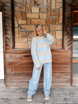 A woman stands on a wooden porch in front of a stone wall, wearing a light blue and beige striped knit sweater, light-wash wide-leg jeans, and beige slip-on shoes.
