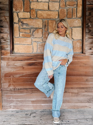A woman stands on a wooden porch in front of a stone wall, wearing a light blue and beige striped knit sweater, light-wash wide-leg jeans, and beige slip-on shoes.