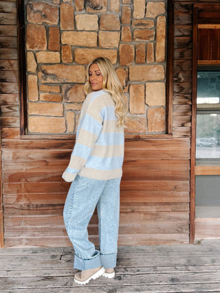 A woman stands on a wooden porch in front of a stone wall, wearing a light blue and beige striped knit sweater, light-wash wide-leg jeans, and beige slip-on shoes.