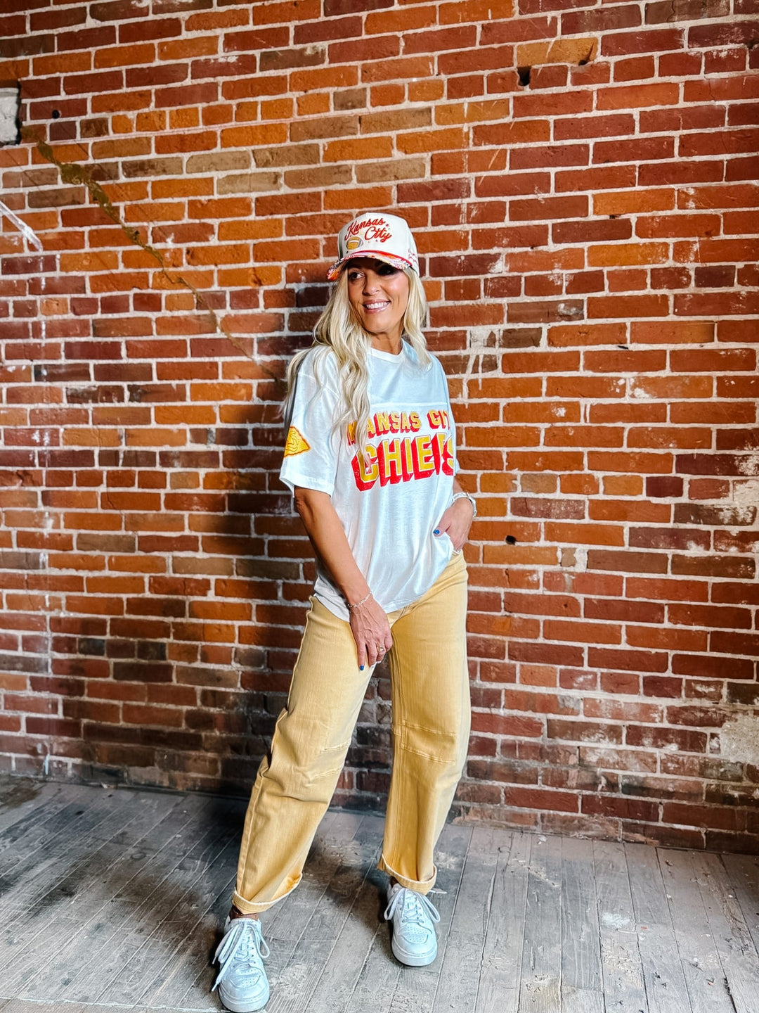 A woman stands in front of a red brick wall wearing a white graphic t-shirt,  gold barrel jeans, a white and red baseball cap, and white sneakers.