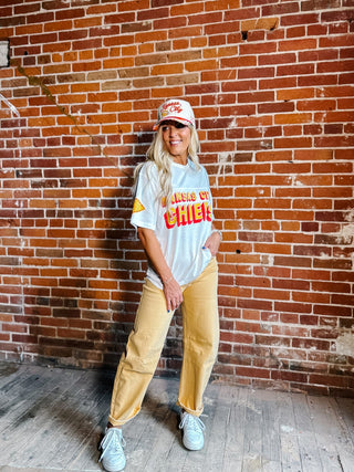 A woman stands in front of a red brick wall wearing a white graphic t-shirt,  gold barrel jeans, a white and red baseball cap, and white sneakers.
