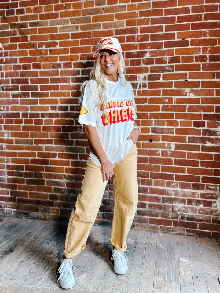 A woman stands in front of a red brick wall wearing a white graphic t-shirt,  gold barrel jeans, a white and red baseball cap, and white sneakers.