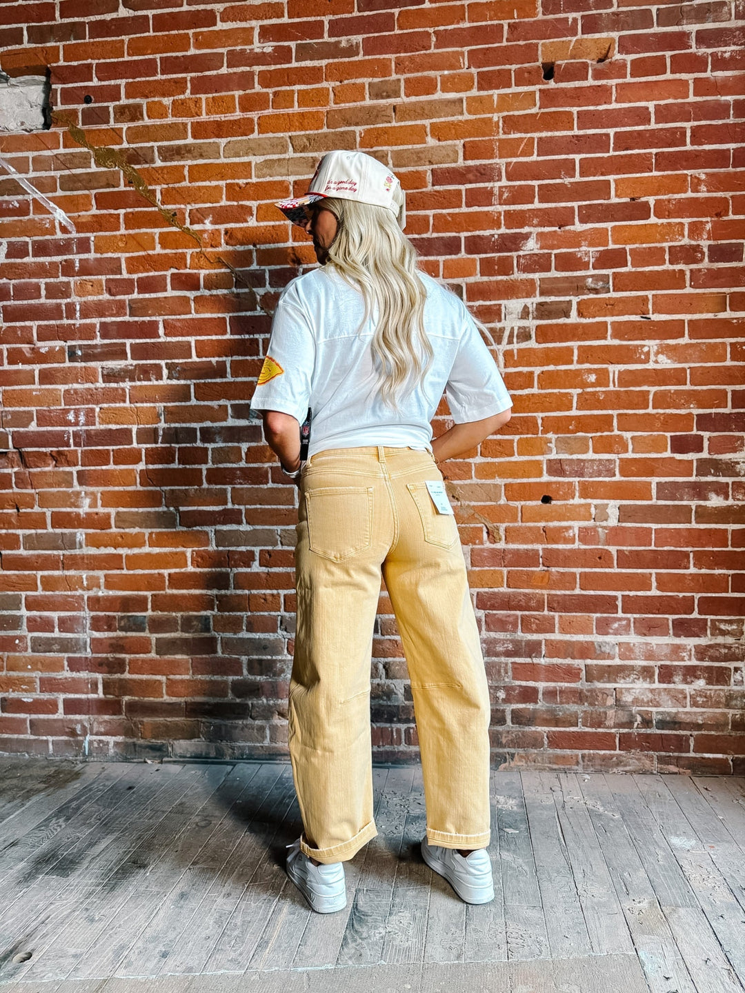 A woman stands in front of a red brick wall wearing a white graphic t-shirt,  gold barrel jeans, a white and red baseball cap, and white sneakers.