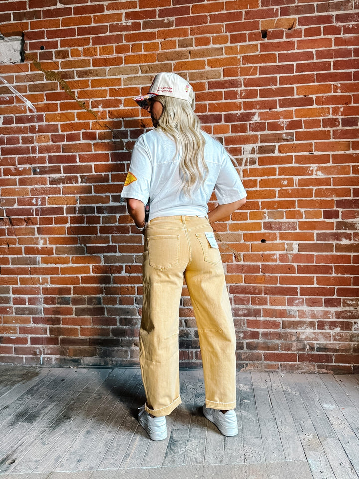 A woman stands in front of a red brick wall wearing a white graphic t-shirt,  gold barrel jeans, a white and red baseball cap, and white sneakers.