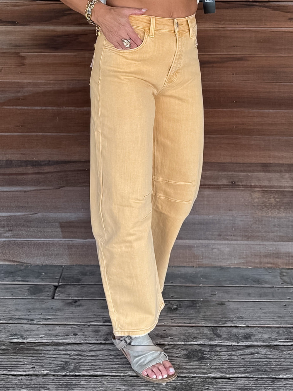 A person wearing wide-leg mustard yellow denim jeans and gray slide sandals, standing on a weathered wooden deck in front of a wooden wall.