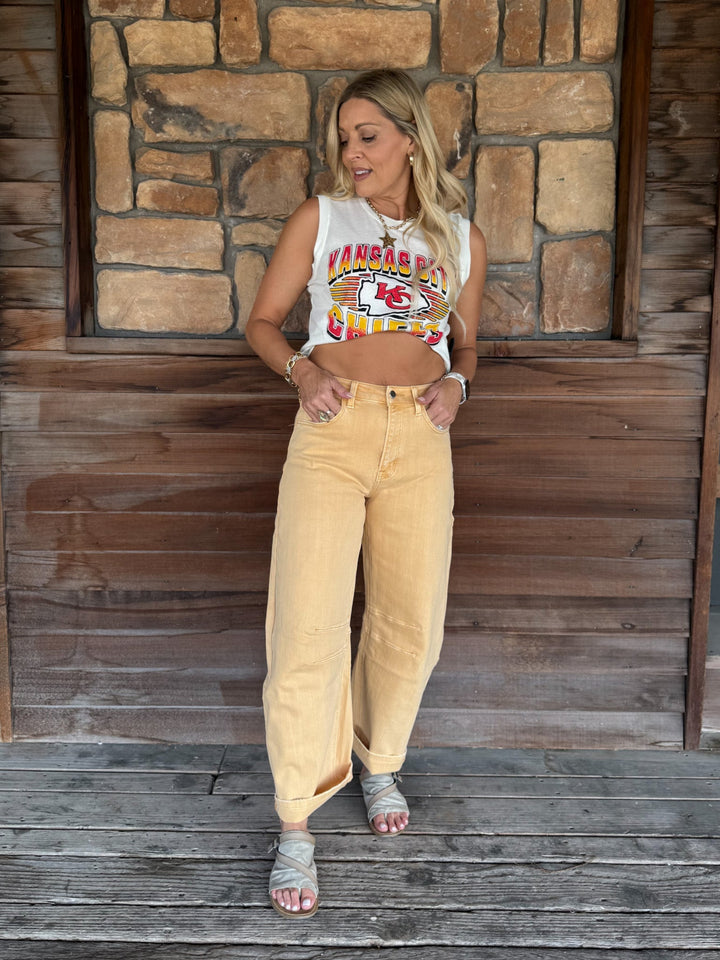 A woman stands on a rustic wooden porch in front of a stone wall, wearing a white sleeveless crop top with a colorful graphic and loose beige wide-leg jeans.