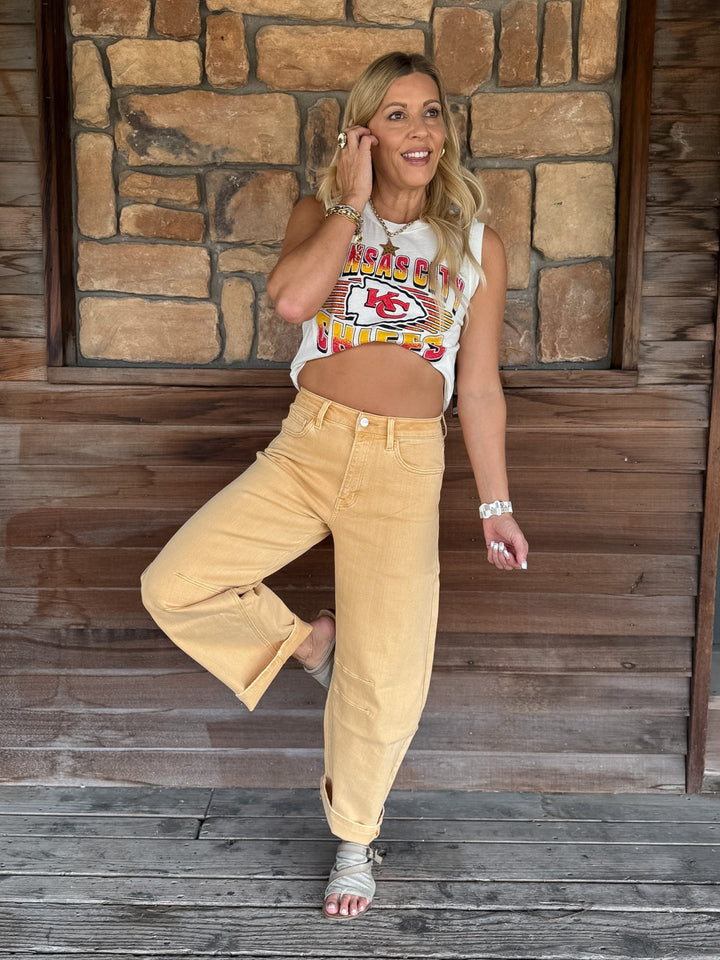 A woman posing on a rustic wooden porch in front of a stone wall, wearing a white sleeveless crop top with a colorful Kansas City Chiefs graphic and light tan wide-leg jeans.
