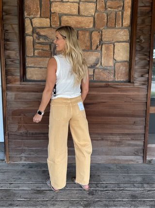 A woman stands on a wooden porch facing away from the camera, wearing wide-leg tan corduroy pants, a white sleeveless top, and pink sandals. The background features a rustic stone wall and wooden door.