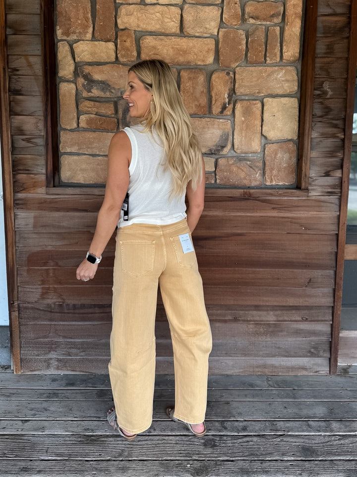 A woman stands on a wooden porch facing away from the camera, wearing wide-leg tan corduroy pants, a white sleeveless top, and pink sandals. The background features a rustic stone wall and wooden door.