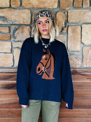 A woman wearing a navy blue knit sweater with a brown horse head graphic on the front, paired with olive green pants and a leopard print headband, standing in front of a rustic stone wall.