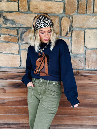 A woman wearing a navy blue knit sweater with a brown horse graphic, olive green pants, a leopard print headband, and a silver chain necklace, standing in front of a rustic stone wall.