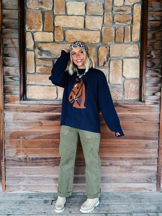 A woman stands in front of a rustic stone and wood wall wearing a navy blue knit sweater with a brown horse graphic, olive green pants, beige boots, and a leopard-print headband.