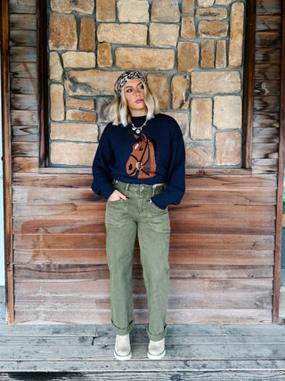 A woman stands on a rustic wooden porch in front of a stone wall, wearing a navy blue crewneck sweatshirt with a brown horse graphic, olive green jeans, and beige boots.