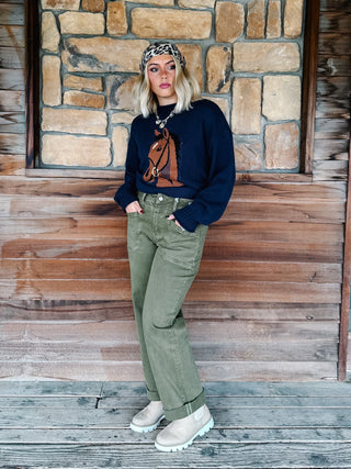 A woman stands on a rustic wooden porch in front of a stone wall, wearing a navy blue long-sleeve top with a brown graphic print, olive green wide-leg jeans, beige boots, and a leopard-print headband.