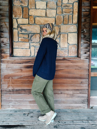 A woman stands on a rustic wooden porch in front of a stone wall, wearing a navy blue knit sweater, olive green pants, and beige boots.