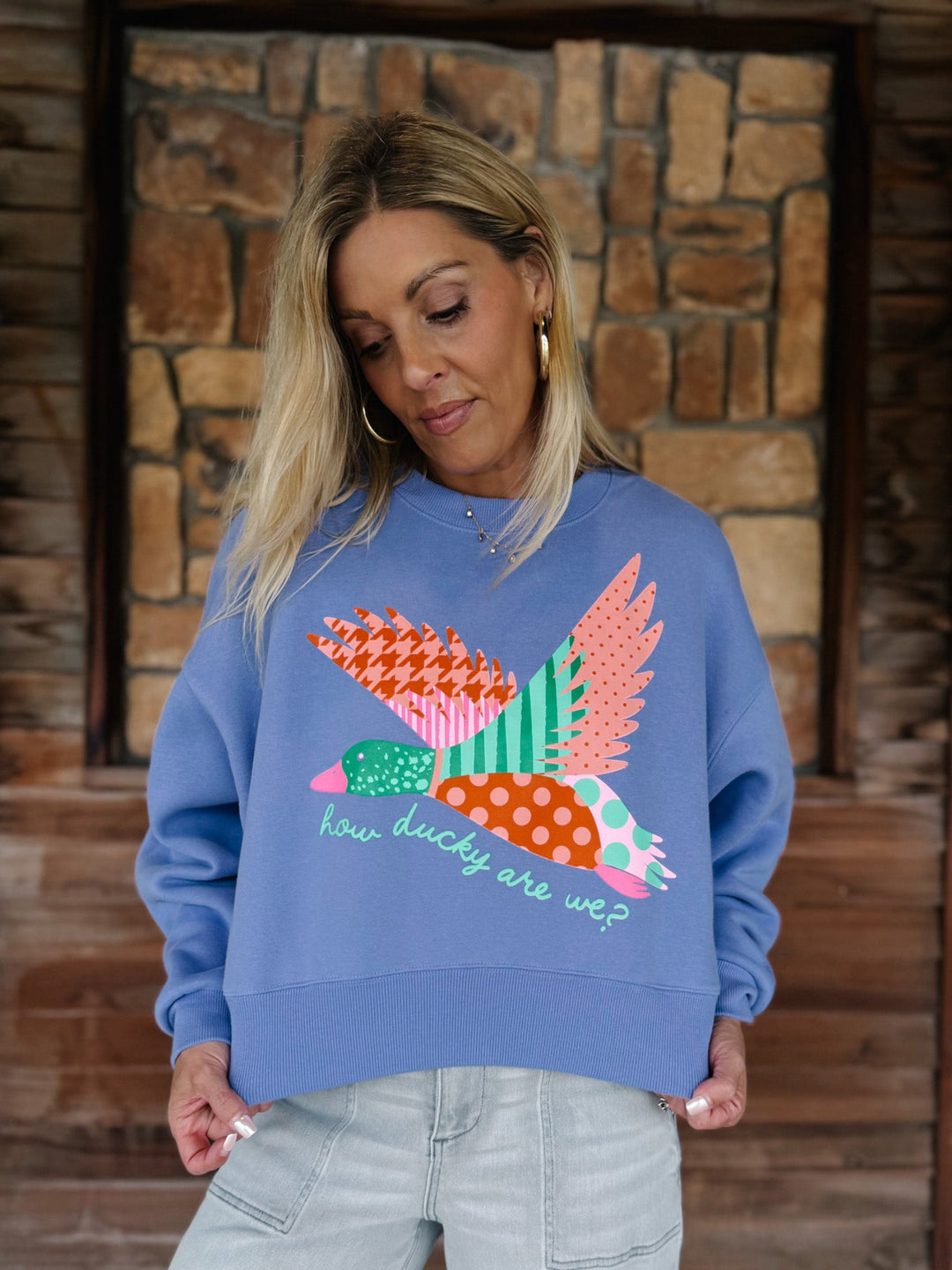A woman wearing a blue crewneck sweatshirt with a colorful patchwork bird graphic and the text 'how ducky are we?' on the front, paired with light gray jeans, standing in front of a rustic stone wall.