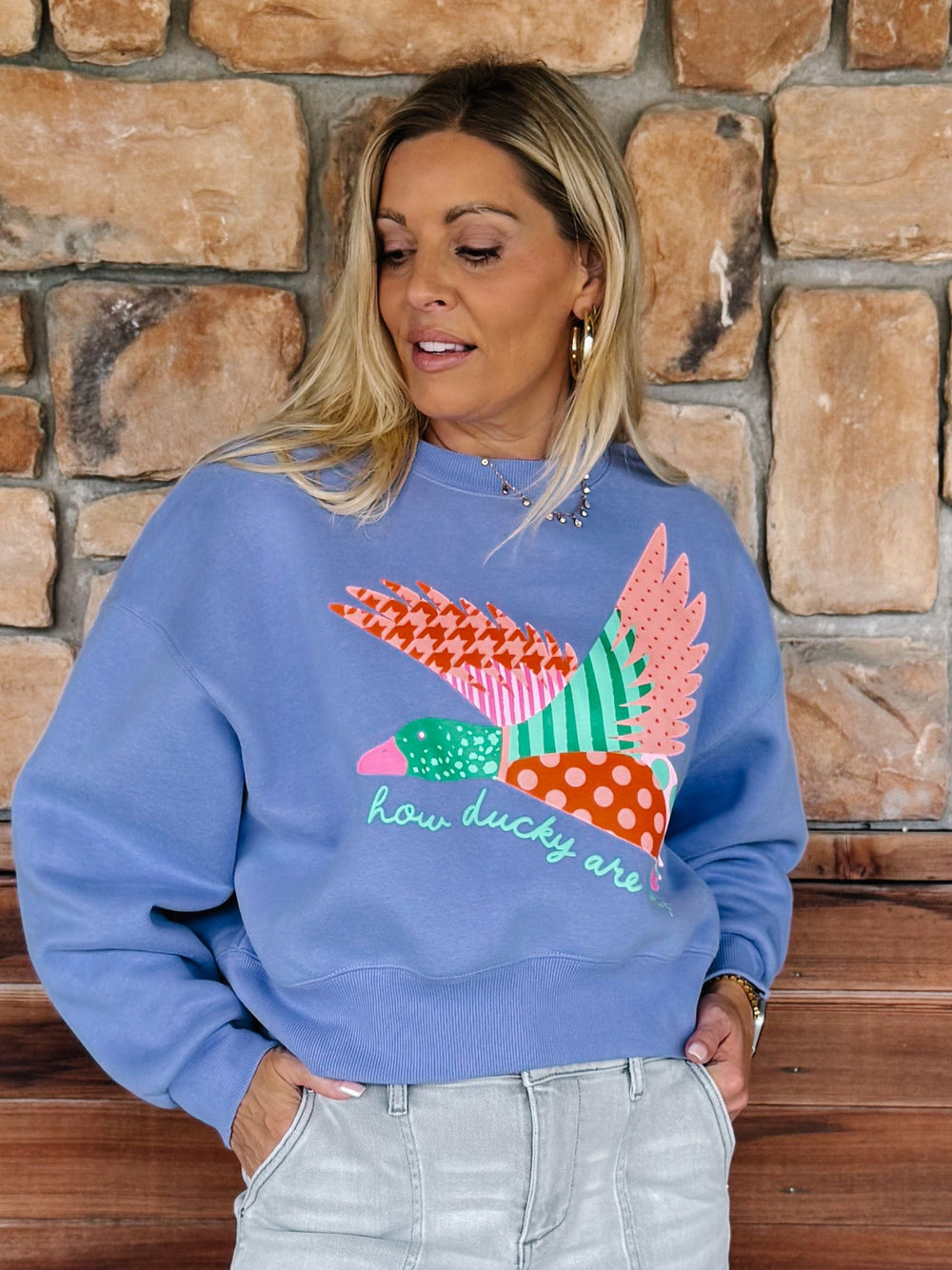 A woman wearing a light blue crewneck sweatshirt with a colorful patchwork duck graphic and the text 'how ducky are you' on the front, paired with light gray jeans, standing in front of a rustic stone wall.