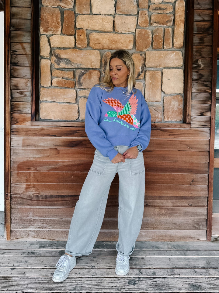 A woman stands on a wooden porch in front of a stone wall, wearing a blue crewneck sweatshirt with a colorful patchwork bird graphic, light-wash wide-leg jeans, and white sneakers.