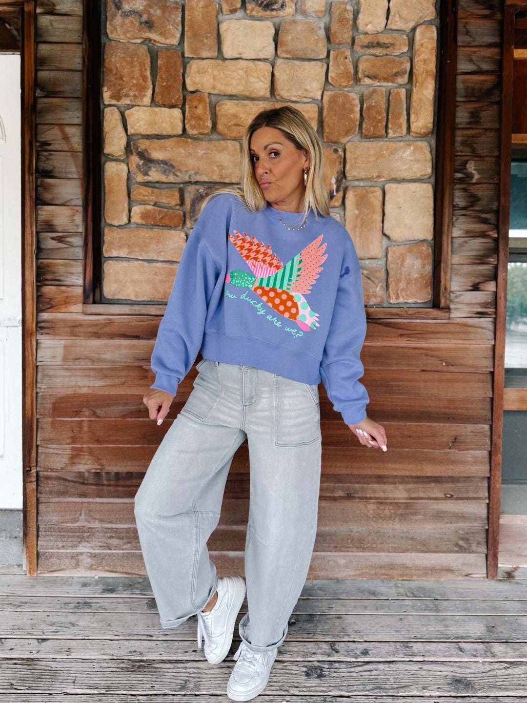 A woman stands on a rustic wooden porch wearing a light blue crewneck sweatshirt with a colorful patchwork bird design, paired with light gray wide-leg jeans and white sneakers.