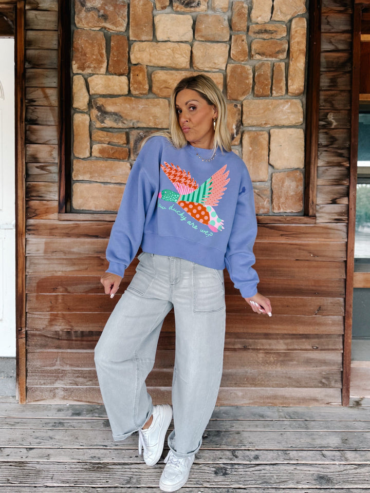 A woman stands on a rustic wooden porch wearing a light blue crewneck sweatshirt with a colorful patchwork bird design, paired with light gray wide-leg jeans and white sneakers.