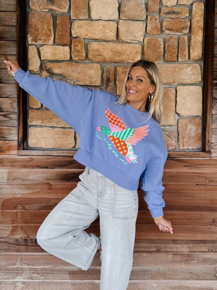 A woman posing in a dusty blue crewneck sweatshirt featuring a colorful patchwork duck on the front, paired with light-wash jeans, set against a rustic stone and wood wall background.