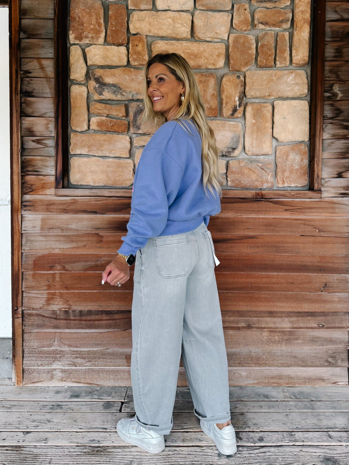 A woman stands on a wooden deck in front of a rustic stone and wood wall, wearing a periwinkle blue sweatshirt, light gray wide-leg jeans, and white sneakers.
