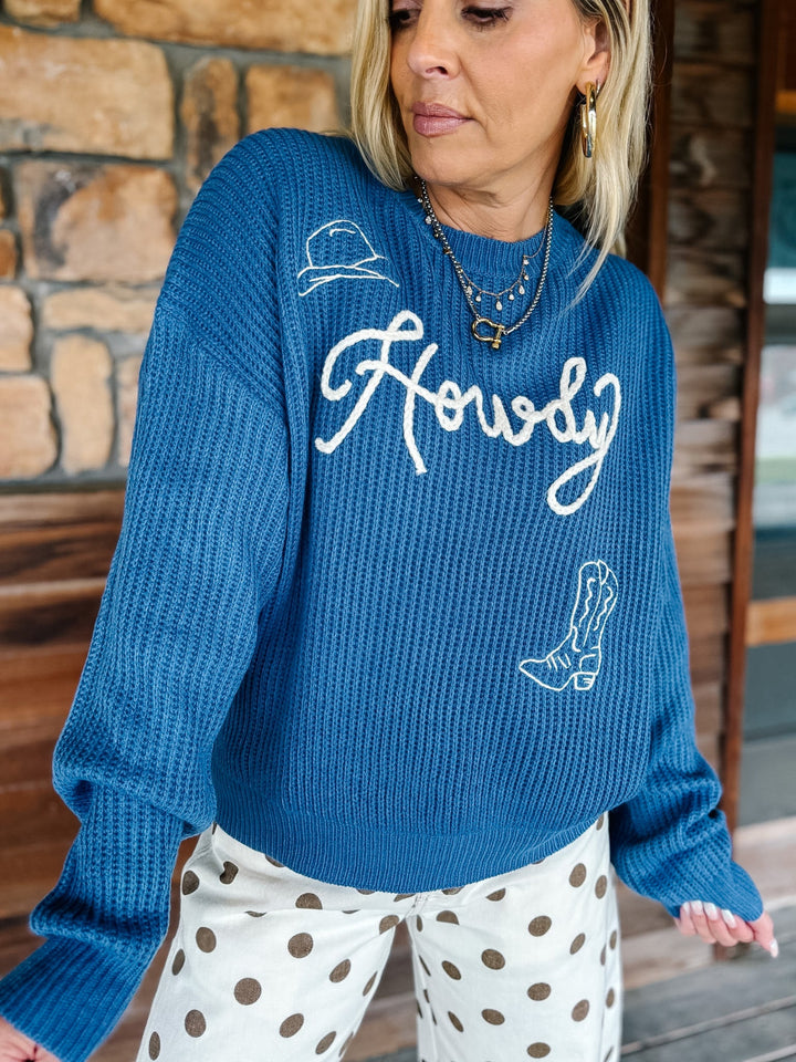 A woman wearing a blue waffle-knit sweater with white embroidered script reading &