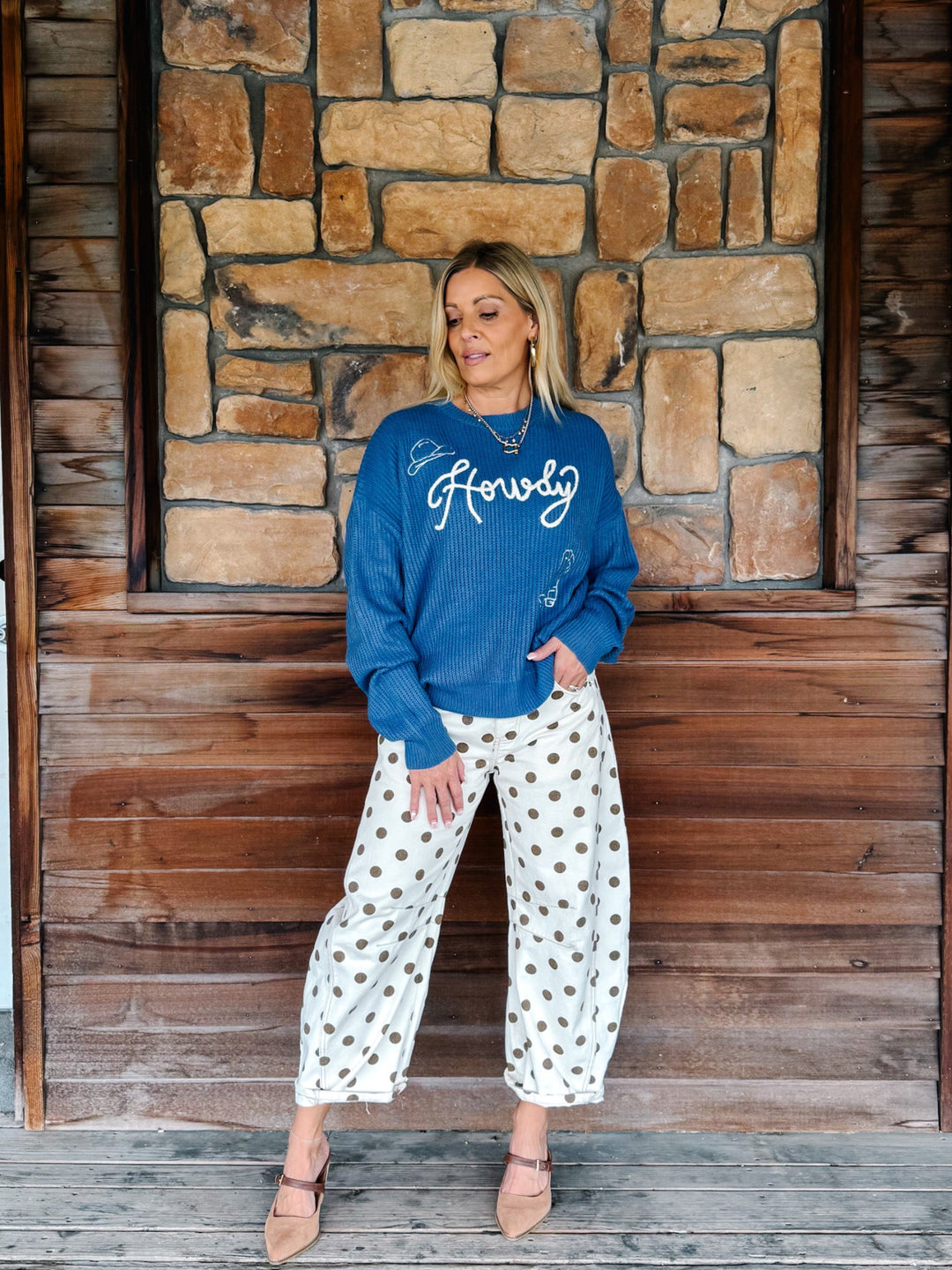 A woman stands in front of a rustic stone and wood wall, wearing a blue crewneck sweatshirt with white script embroidery that reads &