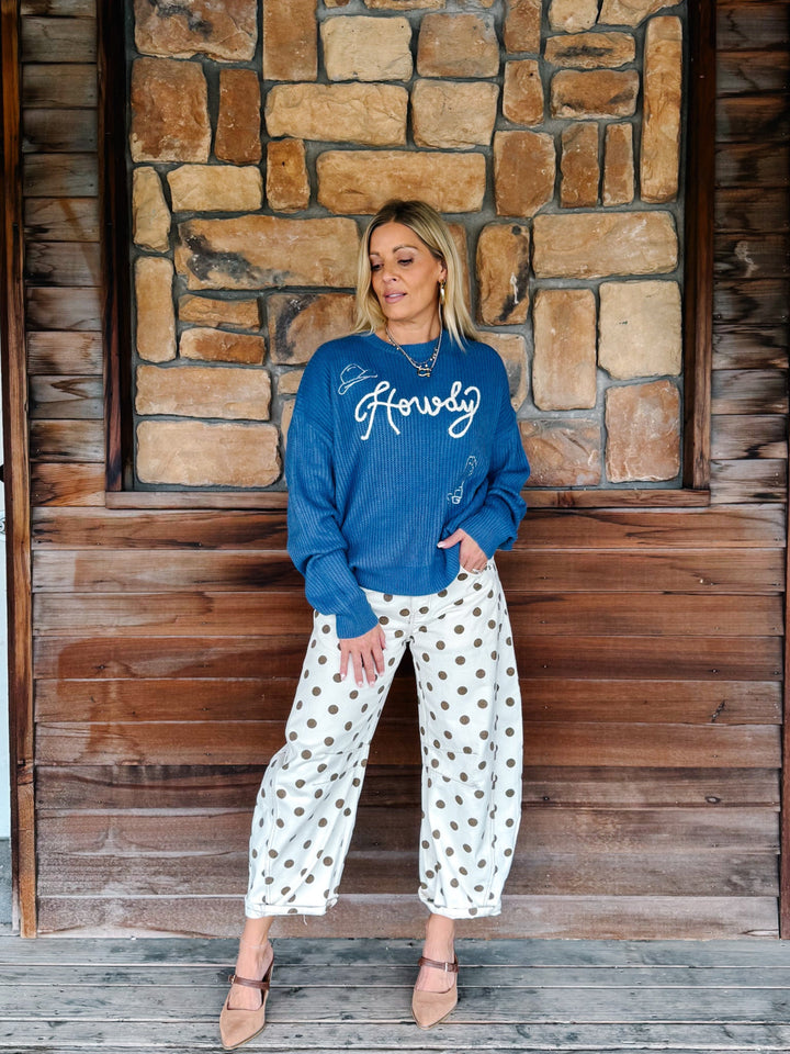 A woman stands in front of a rustic stone and wood wall, wearing a blue crewneck sweatshirt with white script embroidery that reads &