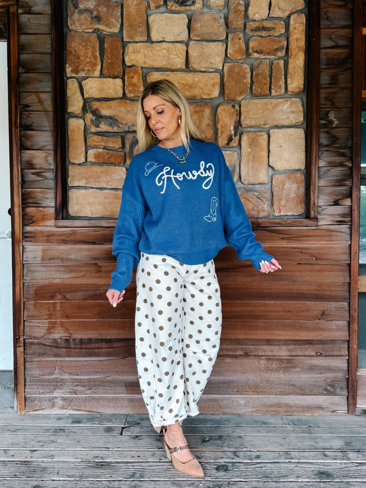 A woman stands on a wooden porch in front of a stone wall, wearing a blue crewneck sweatshirt with &