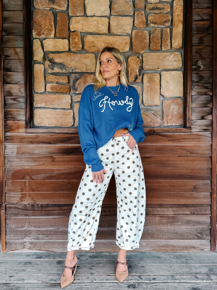 A woman stands against a rustic stone and wood wall wearing a blue crewneck sweatshirt with &