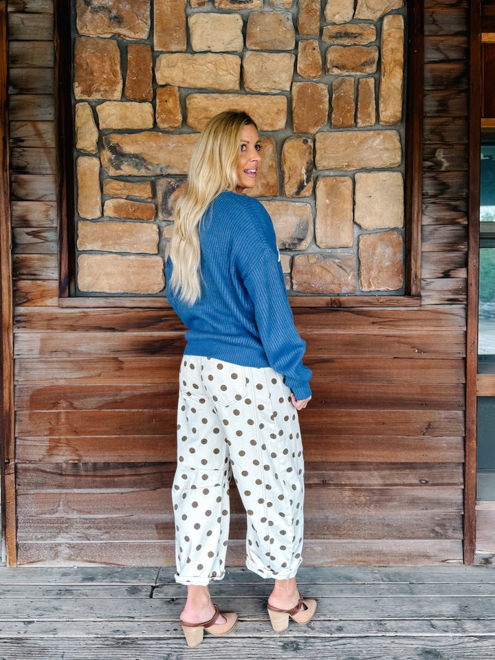 A woman stands on a wooden porch in front of a stone wall, wearing a blue knit sweater and white pants with brown polka dots.