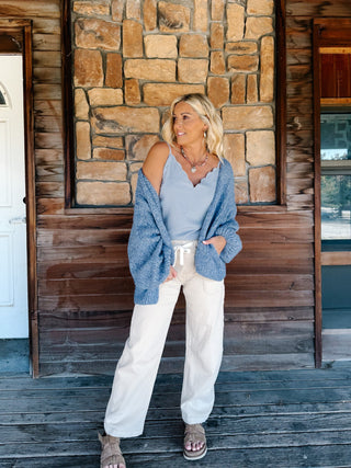 A woman stands on a rustic wooden porch in front of a stone wall, wearing a light blue polka dot tank and a blue knit cardigan, cream  pants, and brown slide sandals.