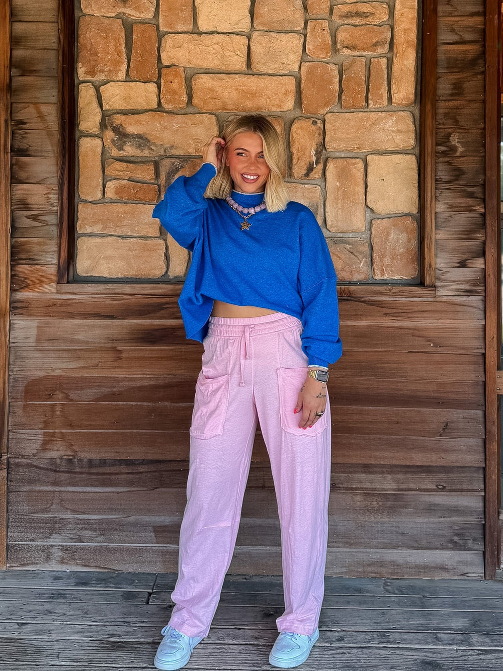 A woman stands on a wooden porch in front of a stone wall, wearing a bright blue cropped long-sleeve top, light pink drawstring pants, and light blue sneakers.