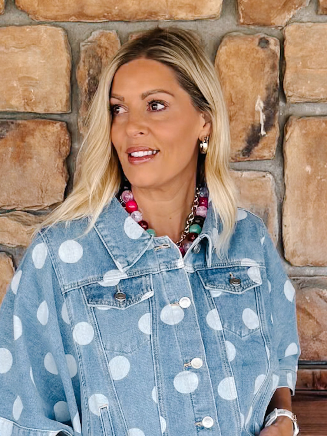 A woman with blonde hair wearing a light blue denim jacket with white polka dots, standing in front of a rustic stone wall.