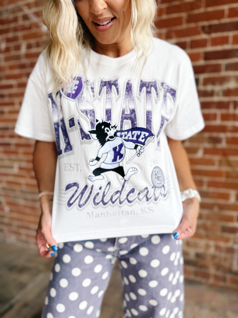 A woman wearing a white graphic t-shirt with a purple and black 'K-State Wildcats' print, paired with gray and white polka dot pants, standing in front of a brick wall.
